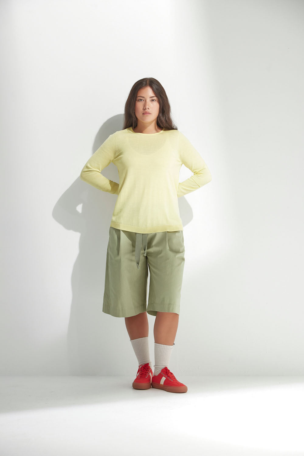 Woman wearing a light yellow long-sleeve top, green shorts, and red shoes on a white background