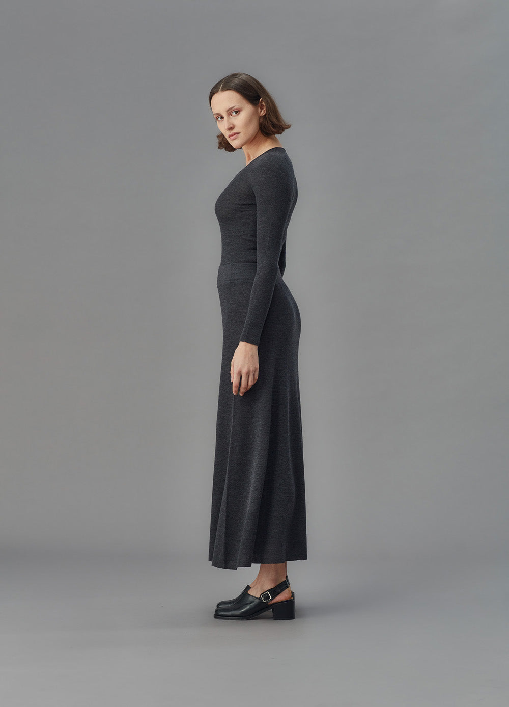 Woman wearing a long dark gray skirt and u top against a gray background