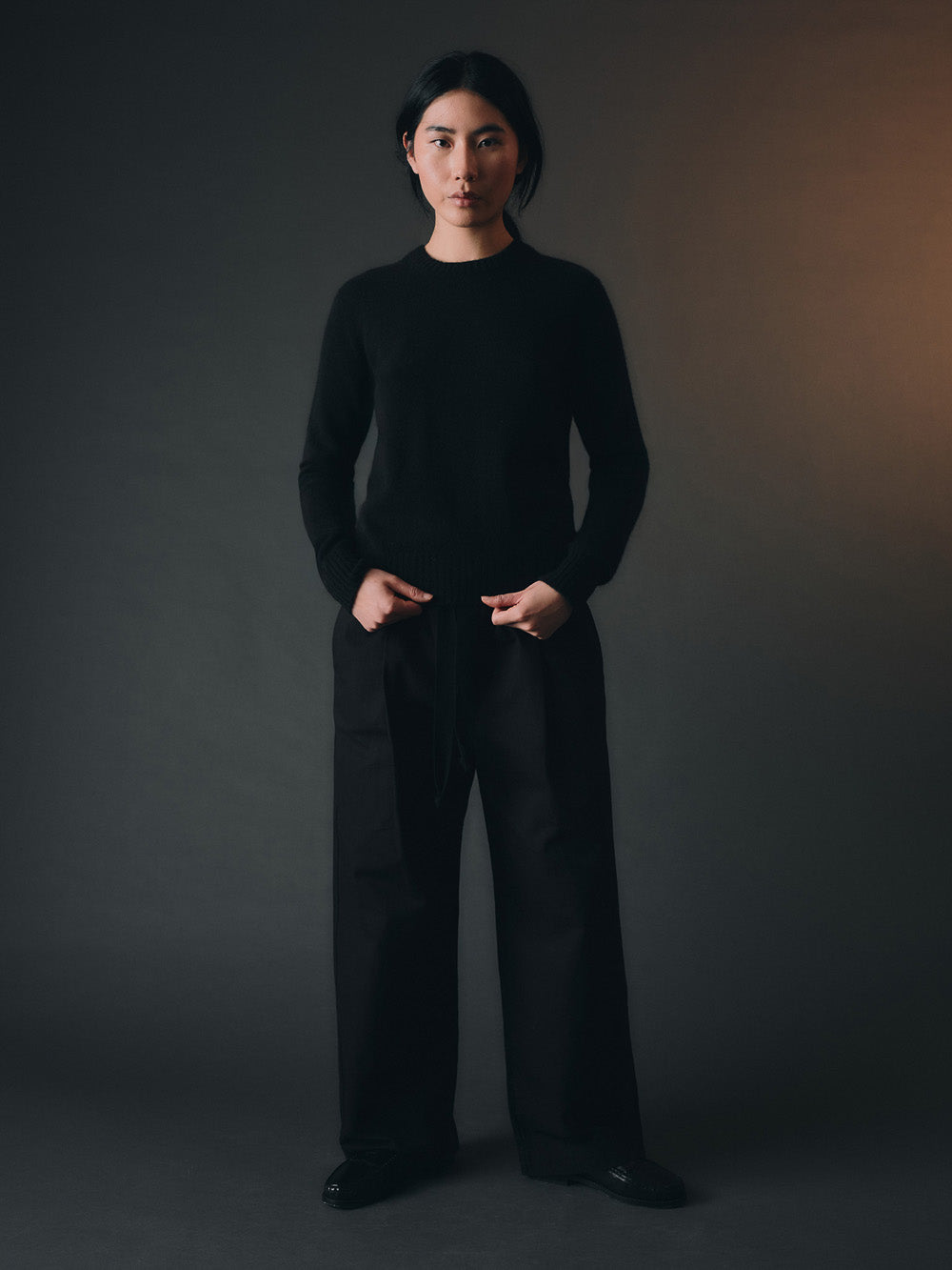 Person wearing a black sweater and trousers against a dark background