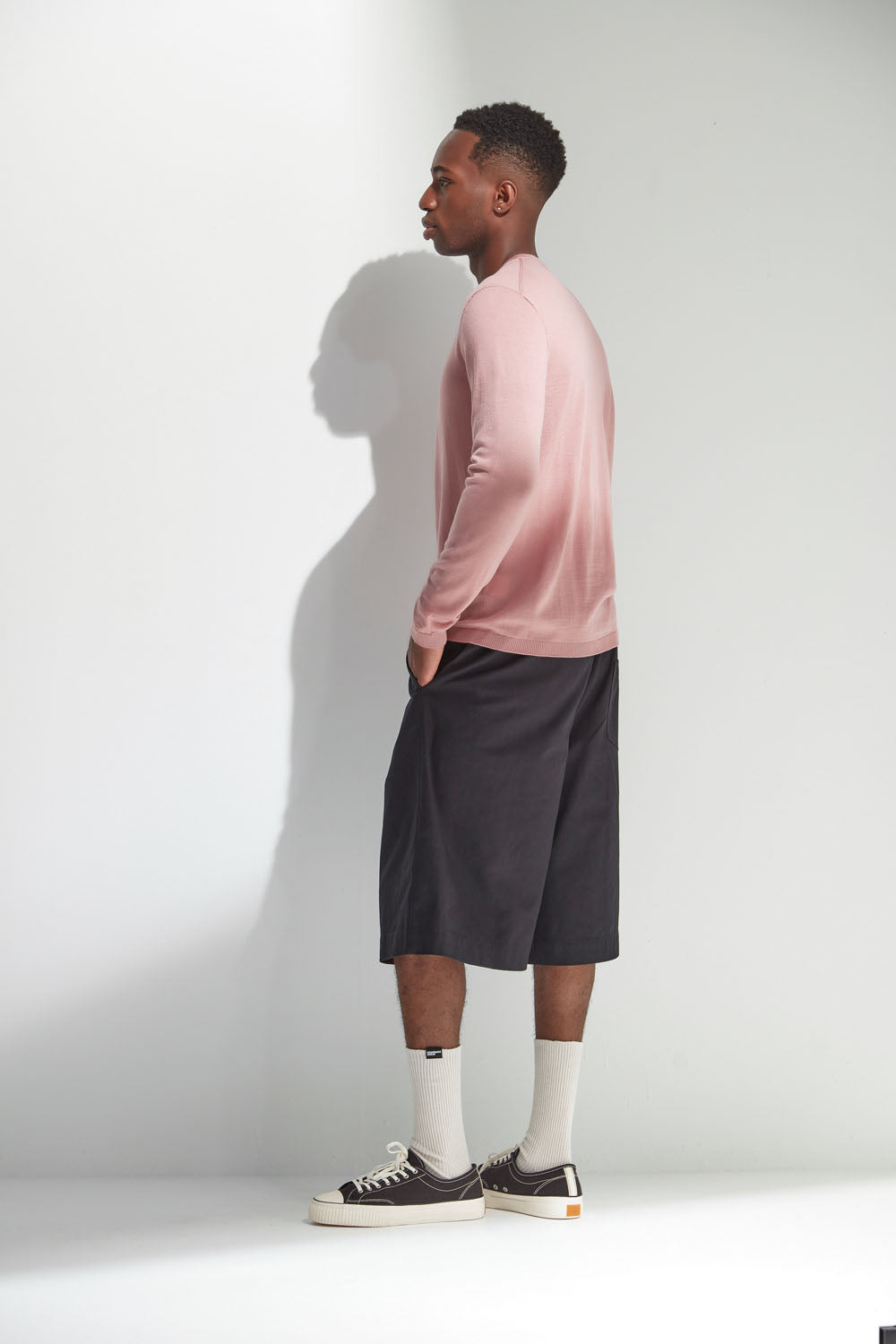 Person wearing a pink long-sleeve shirt and black shorts standing against a white wall.