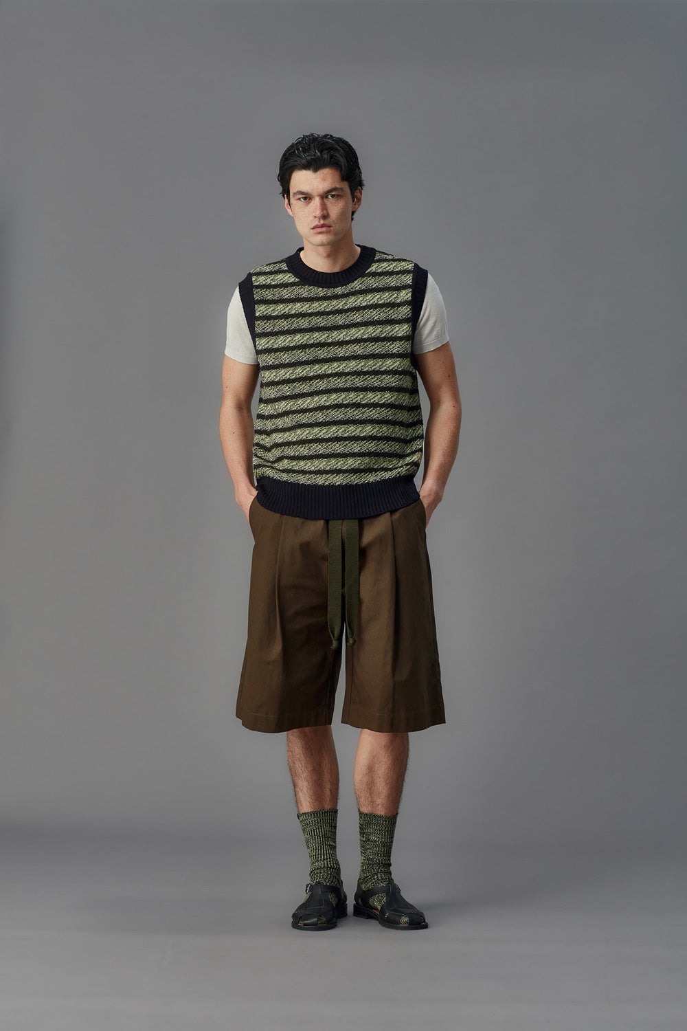 Man wearing a striped vest and white shirt with brown shorts against a gray background