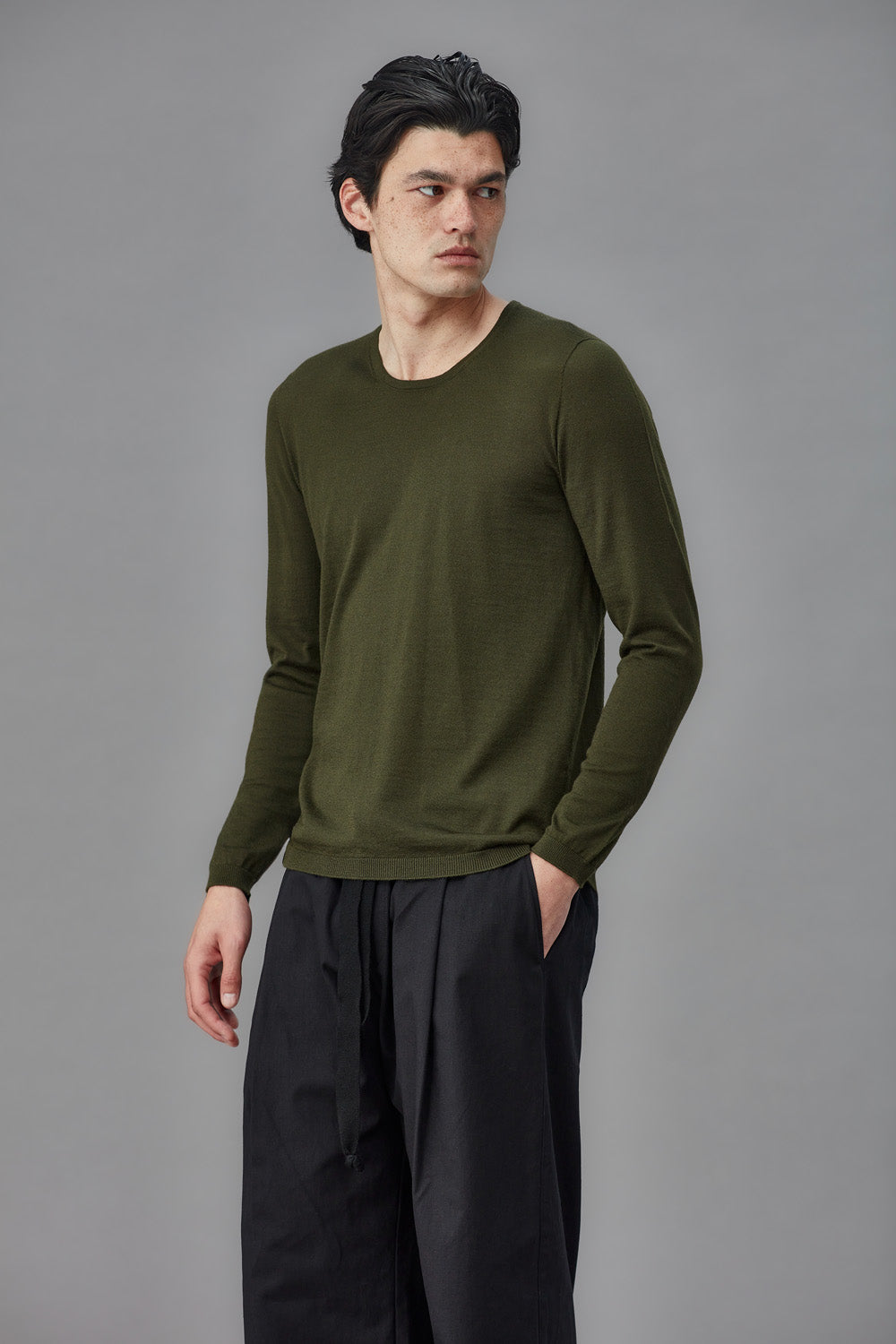 Man wearing an olive green long-sleeve shirt and black pants against a gray background