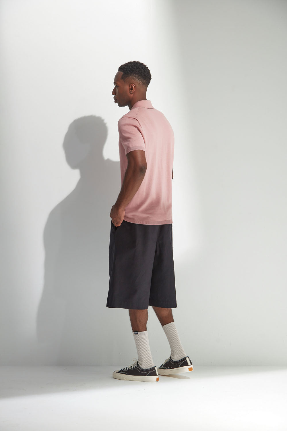 Man wearing a coral polo shirt and black shorts standing against a white wall.