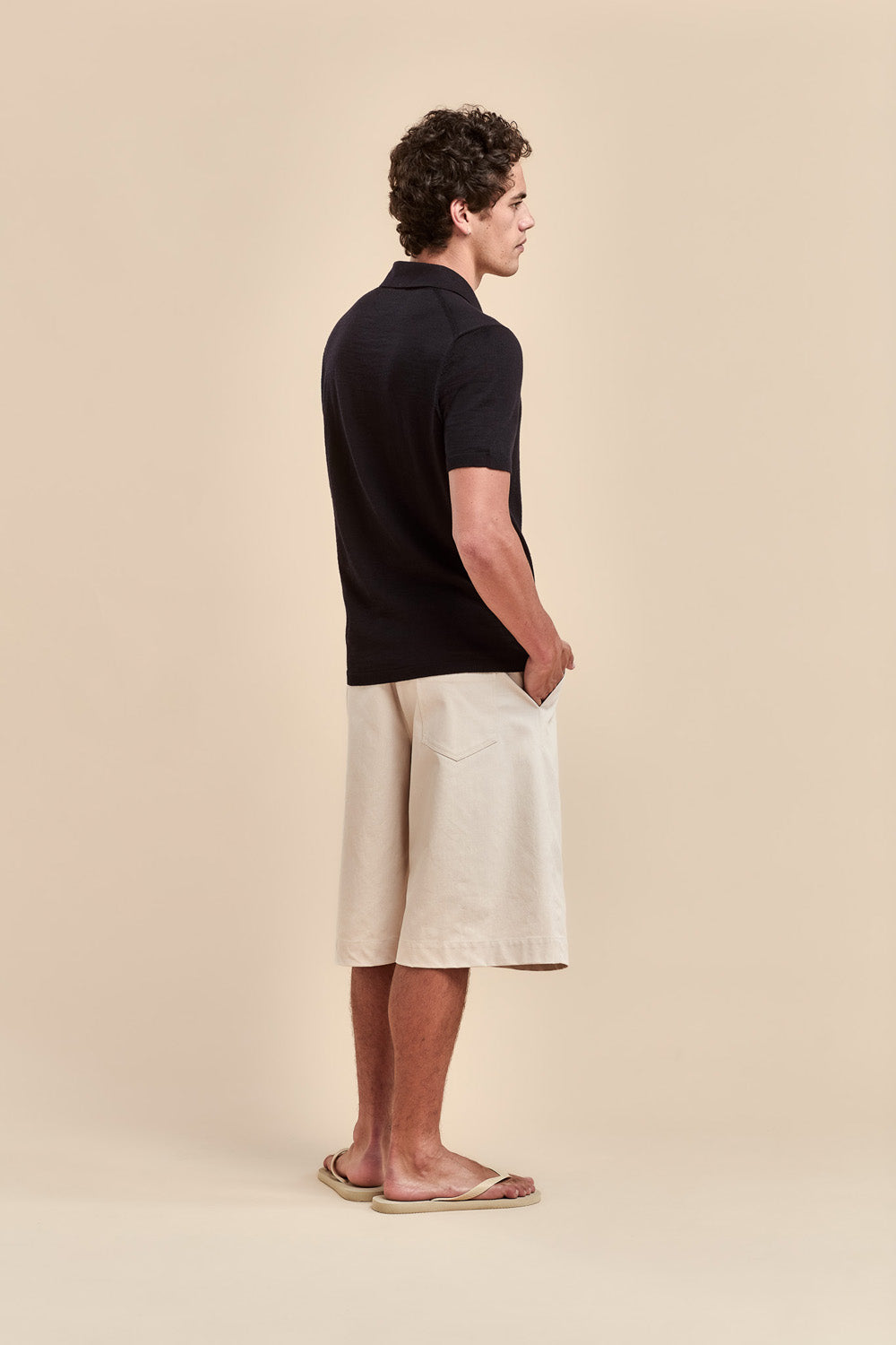 A man stands in profile wearing a black short-sleeve polo shirt, off-white shorts, and tan sandals against a solid beige studio background.