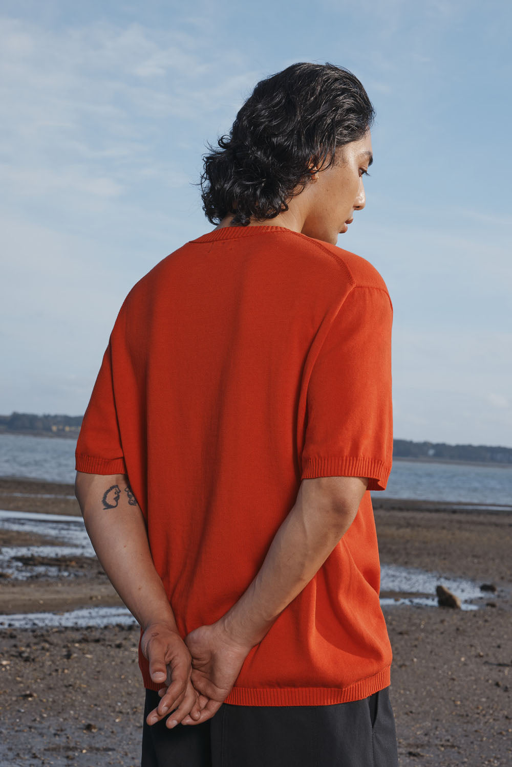 Man wearing a red shirt and black shorts standing on a beach with a blue sky.