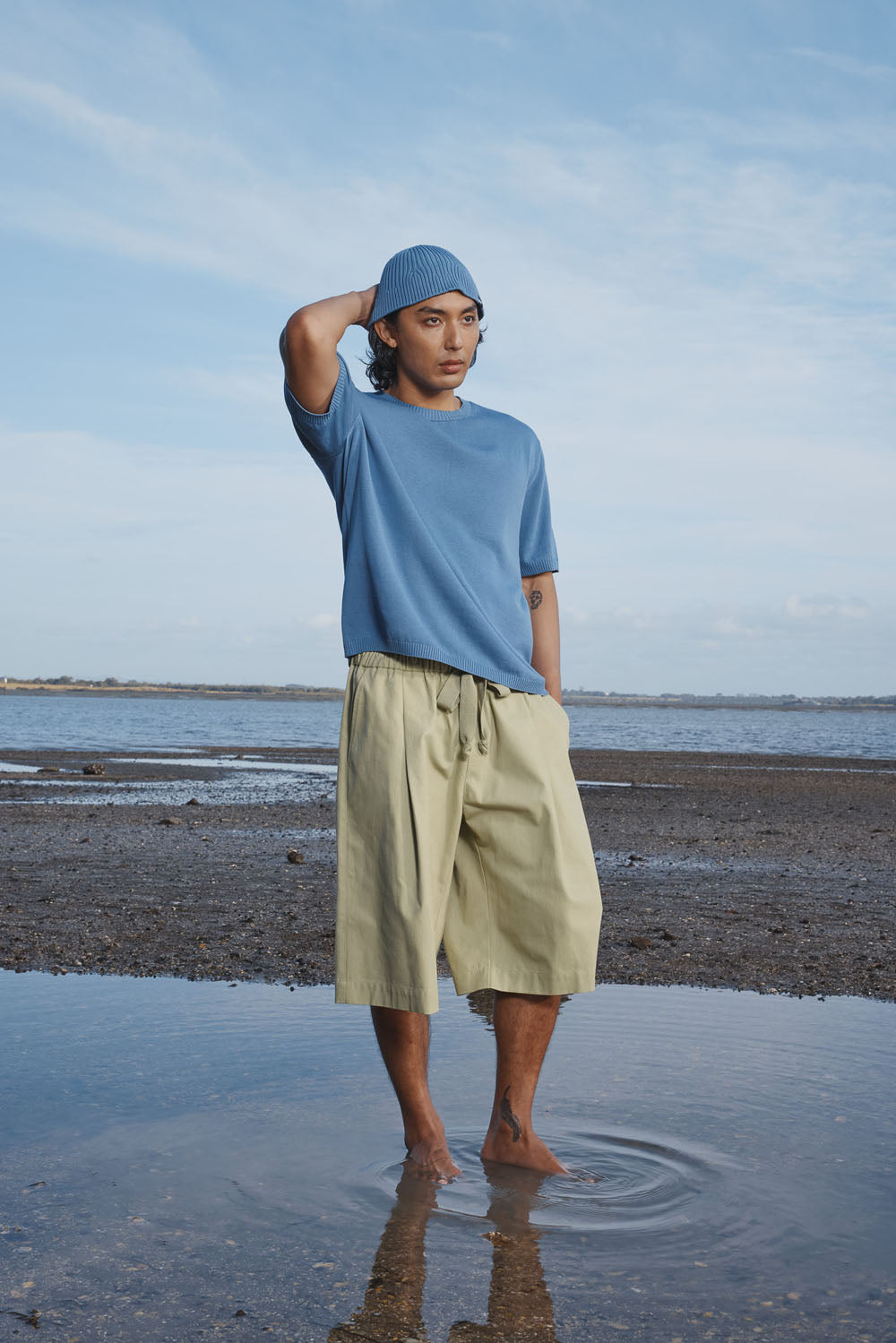 Man standing on a beach wearing a blue shirt and green shorts, with a clear sky in the background.