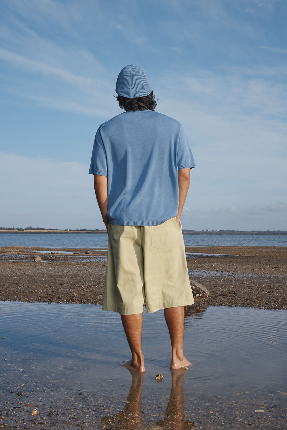 Man standing on a beach wearing a blue shirt and green shorts, with a clear sky in the background.