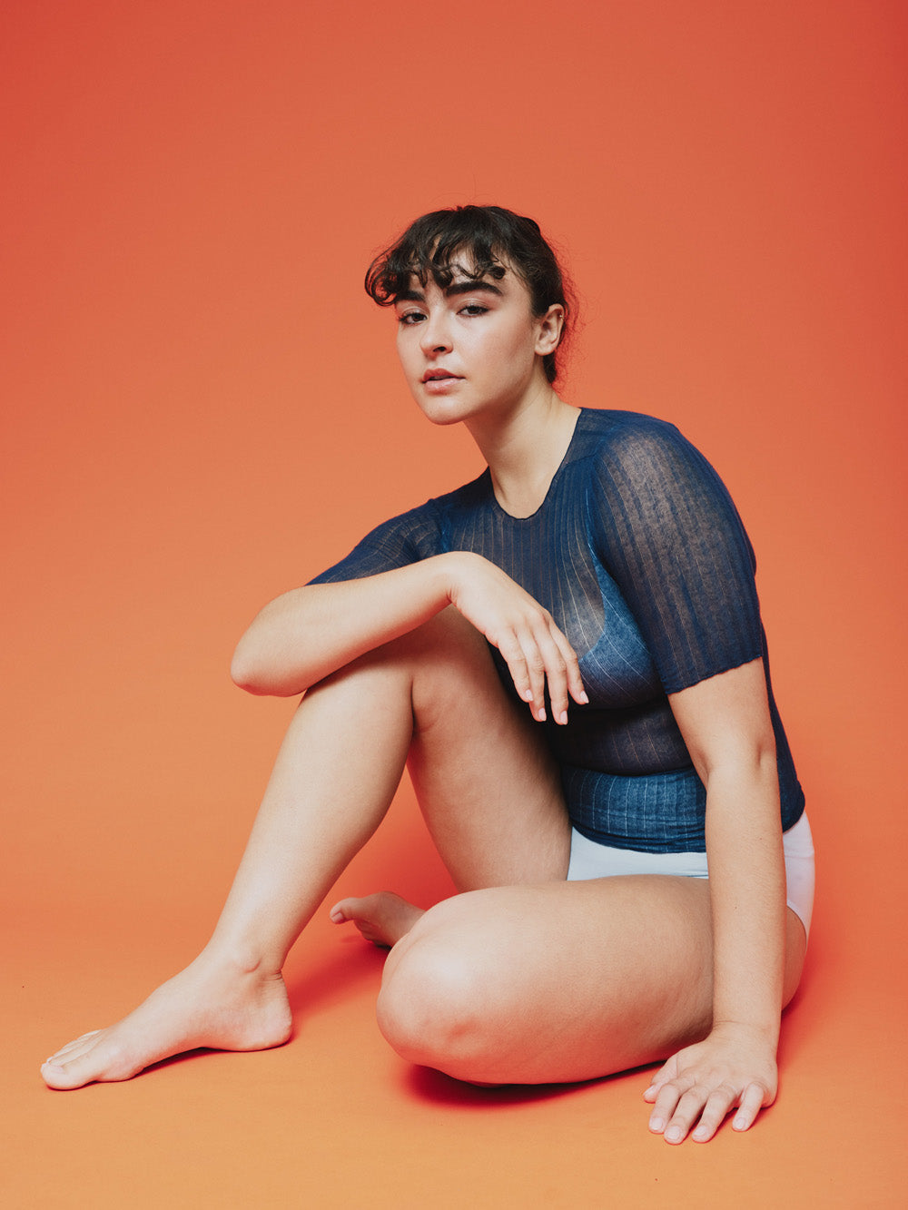 Woman sitting on an orange background wearing a blue sheer tulle top and white underwear.
