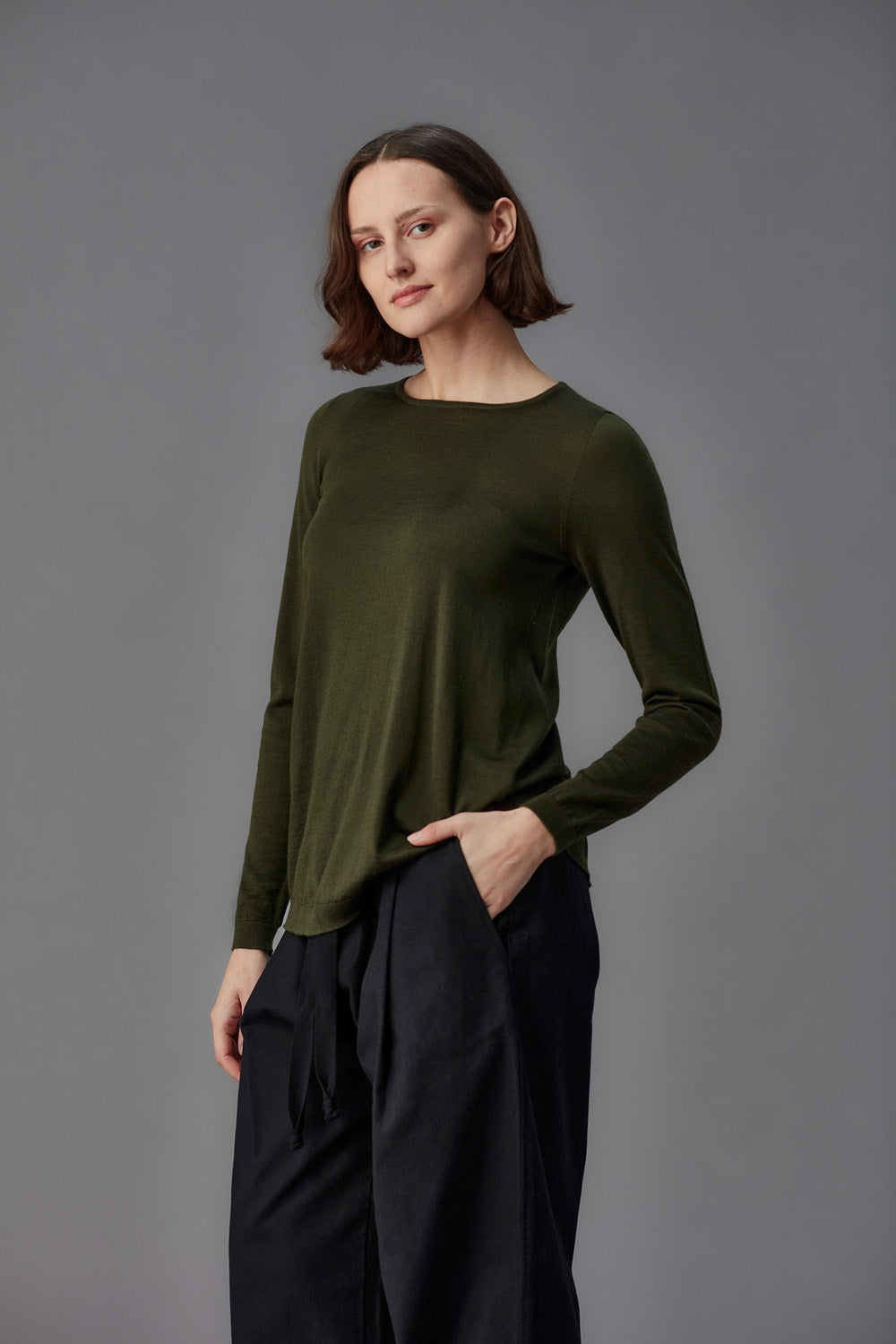 Woman wearing an olive green long-sleeve top and black pants against a gray background
