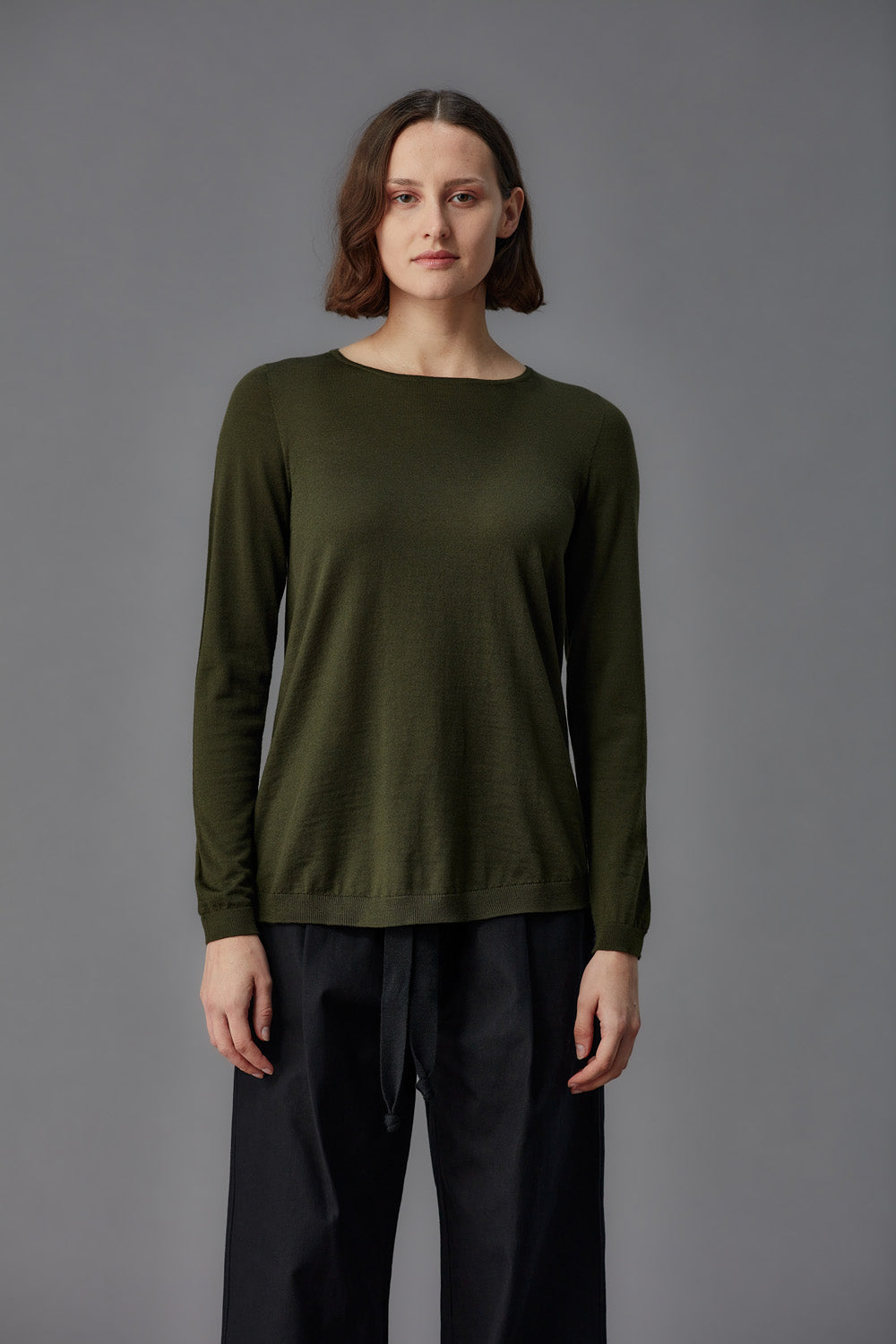 Person wearing a green long-sleeve shirt and black pants against a gray background