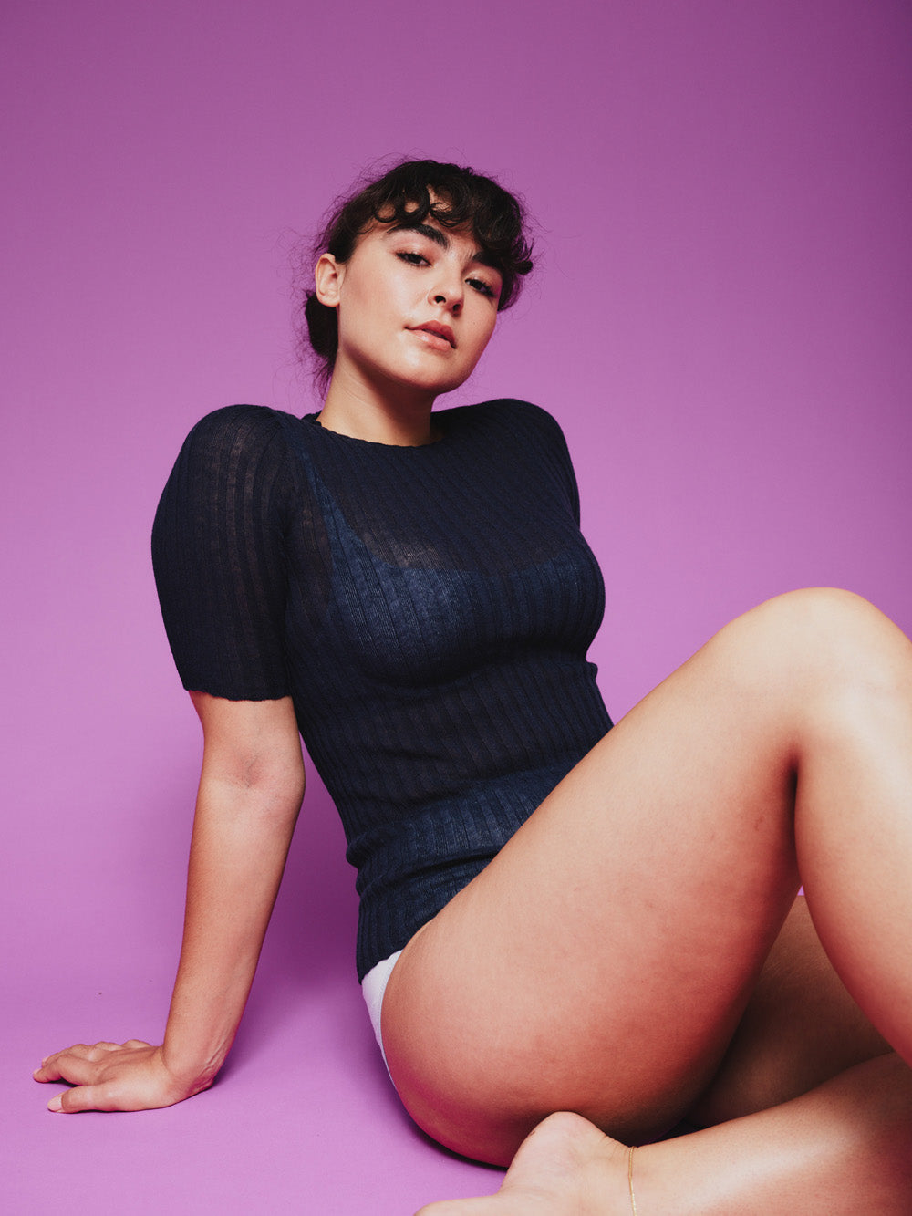 Woman sitting on a purple background wearing a dark blue merino tulle top