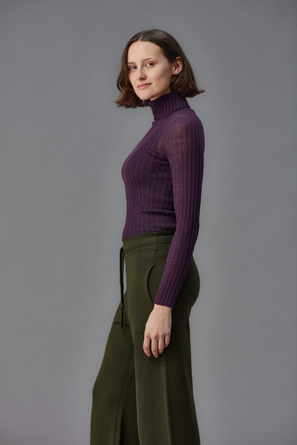 Woman wearing a purple ribbed turtleneck tulle and green pants against a gray background