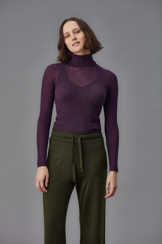 Woman wearing a purple ribbed turtleneck tulle and green pants against a gray background