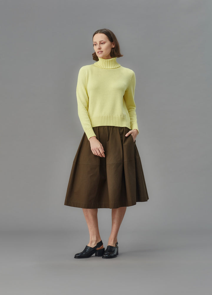 Woman wearing a light yellow funnel sweater and brown skirt against a gray background