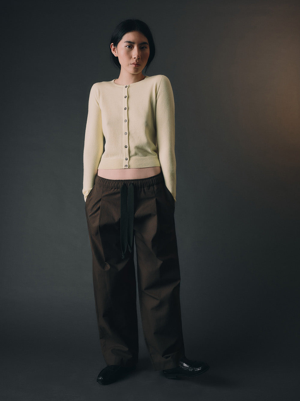 Woman wearing a light yellow cashmere cardigan and brown pants against a dark background