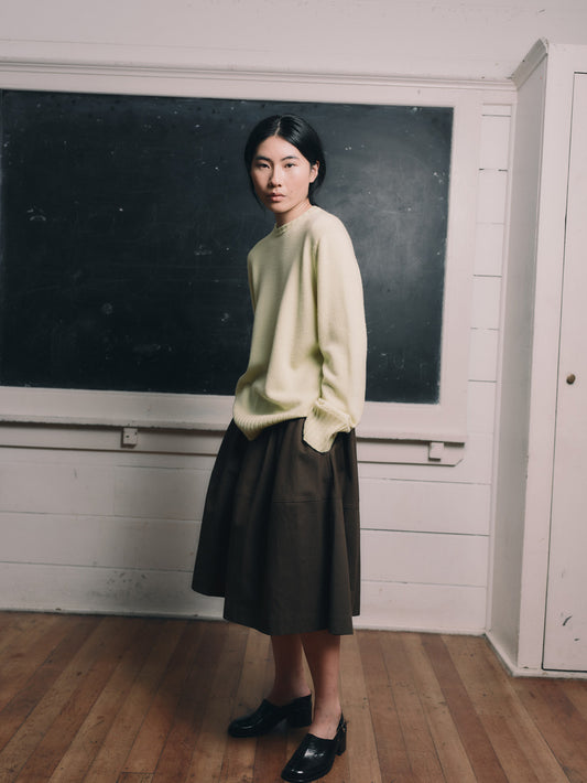 Woman in a light yellow cashmere sweater and dark skirt standing in front of a blackboard.