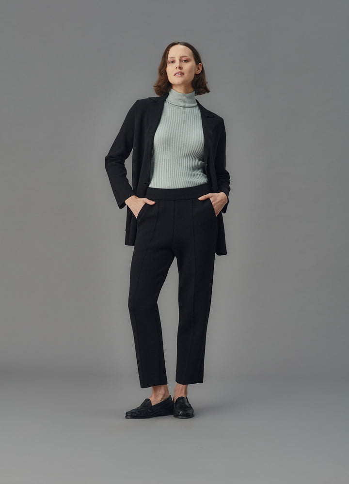 Woman wearing a black merino blazer, light blue turtleneck, and black pants against a gray background
