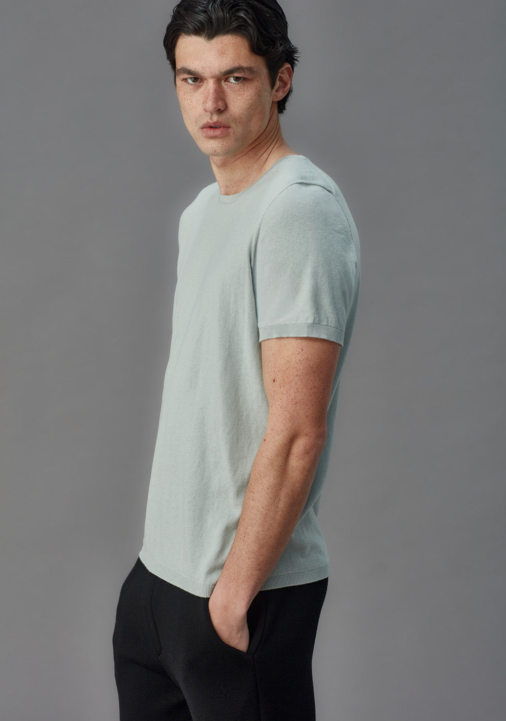 Man wearing a light blue t-shirt and black pants against a gray background