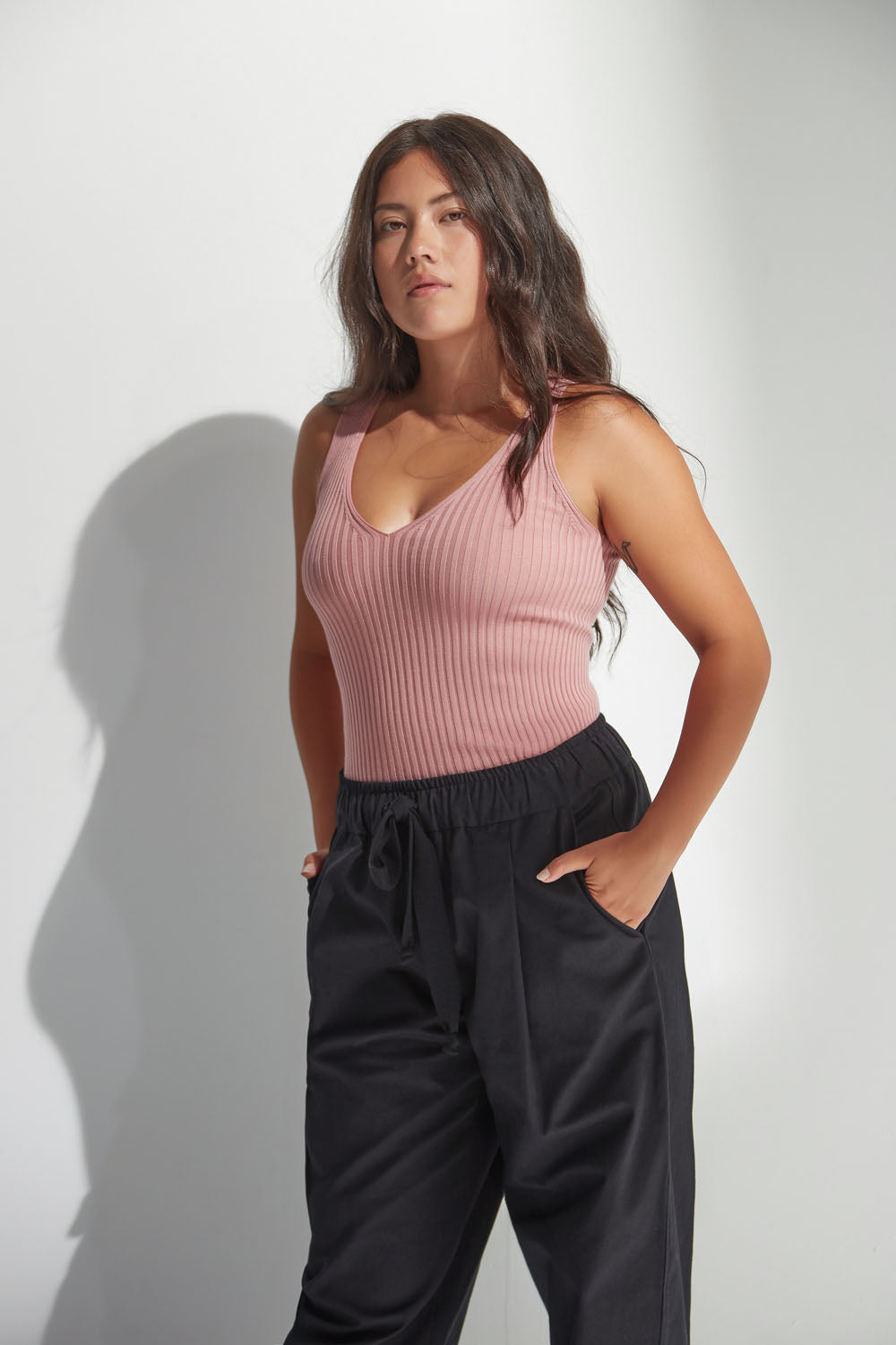 Woman wearing a pink ribbed tank top and black pants against a white background