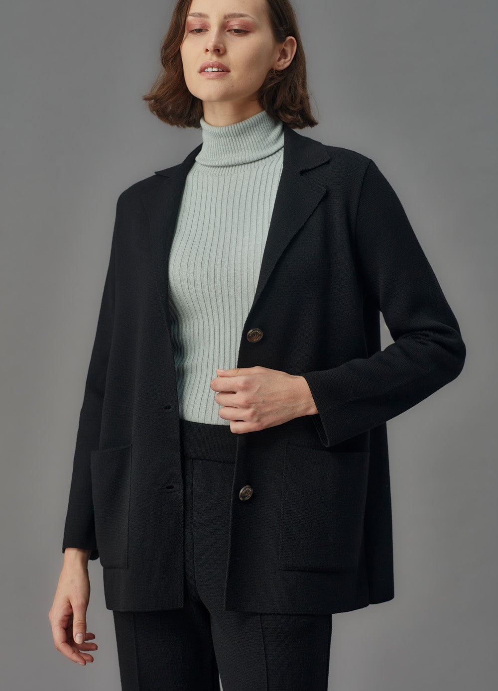 Woman wearing a black merino jacket and pants over a light blue turtleneck on a gray background