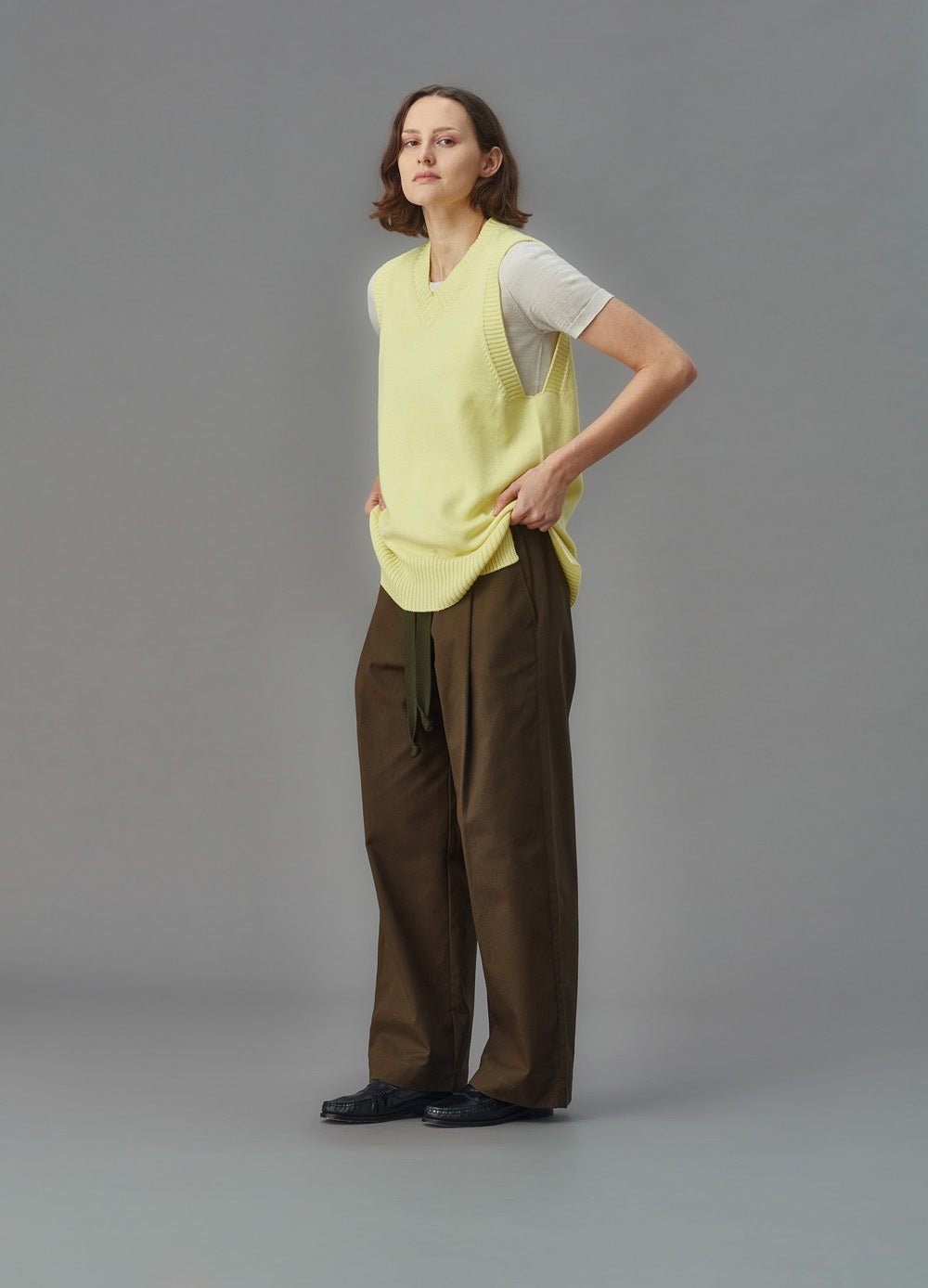 Woman wearing a light yellow sleeveless sweater vest over a white shirt on a gray background