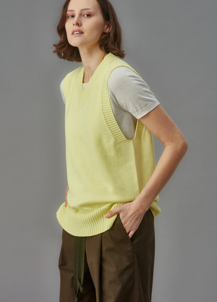 Woman wearing a light yellow sleeveless sweater vest over a white shirt on a gray background