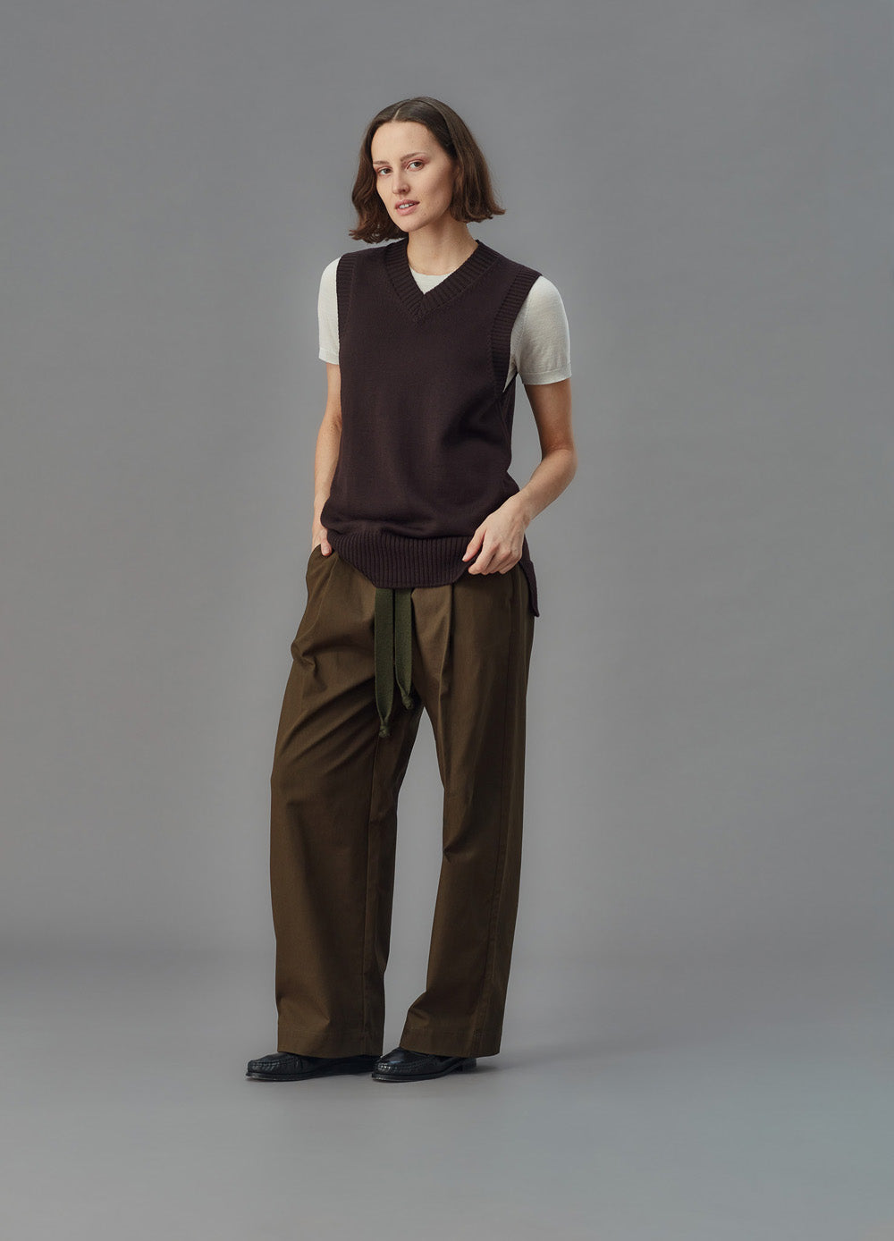 Person wearing a brown sweater vest over a white shirt with brown pants on a gray background