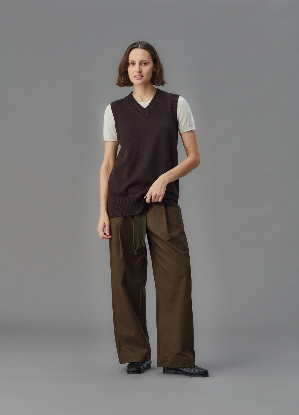 Person wearing a brown sweater vest and pants on a gray background