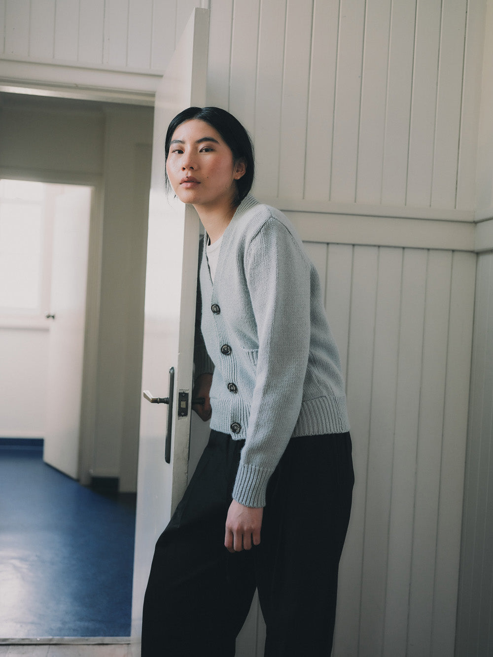 Woman wearing a light blue cardigan and black pants standing in a room with white walls and a blue floor.
