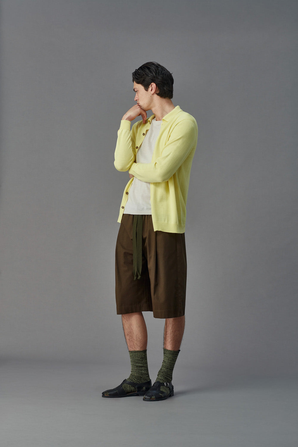 Person wearing a light yellow cardigan over a white shirt with brown shorts on a gray background