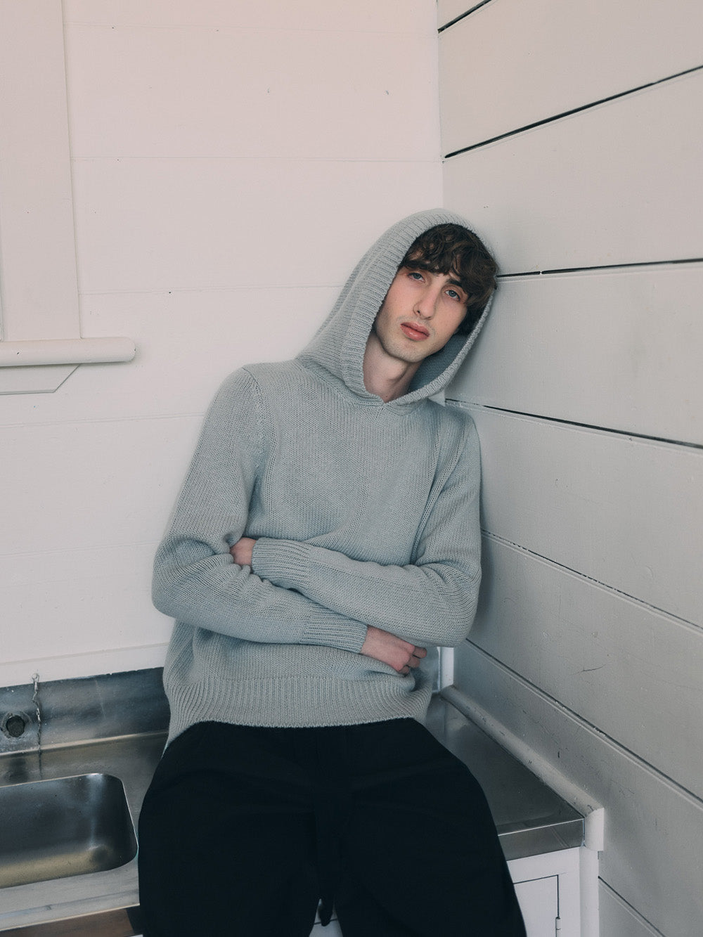 Person wearing a light blue hooded knit sweatshirt sitting on a bench in a minimalistic room.