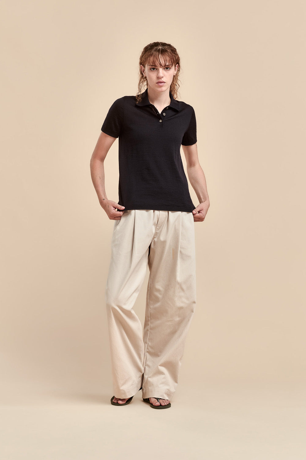 A woman stands against a plain beige studio background wearing a black merino short-sleeve polo shirt and loose beige wide-leg pants.