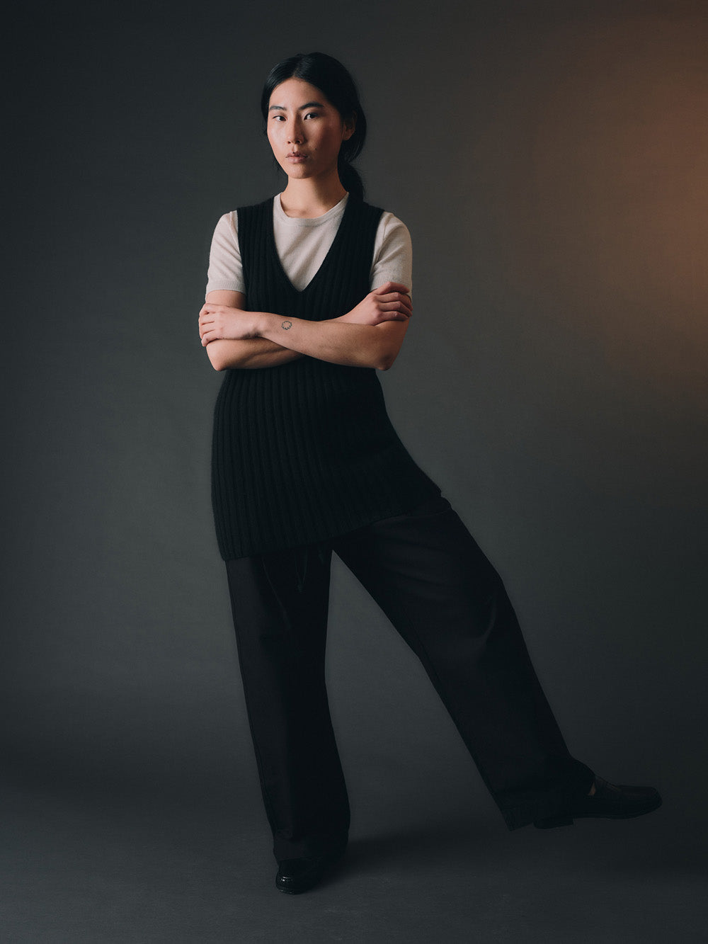 Woman wearing a black knitted vest over a white shirt and black trousers against a dark background.