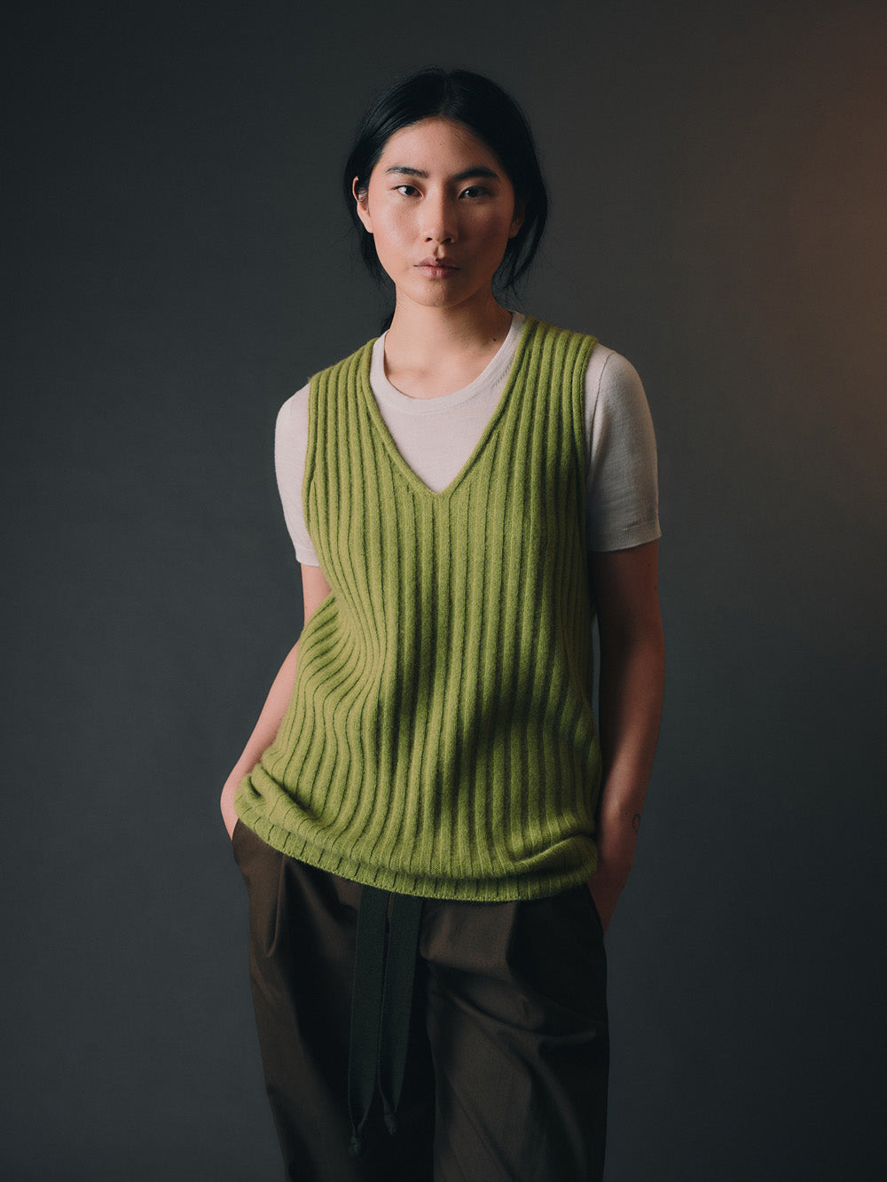 Woman wearing a green knitted vest over a white shirt against a dark background.