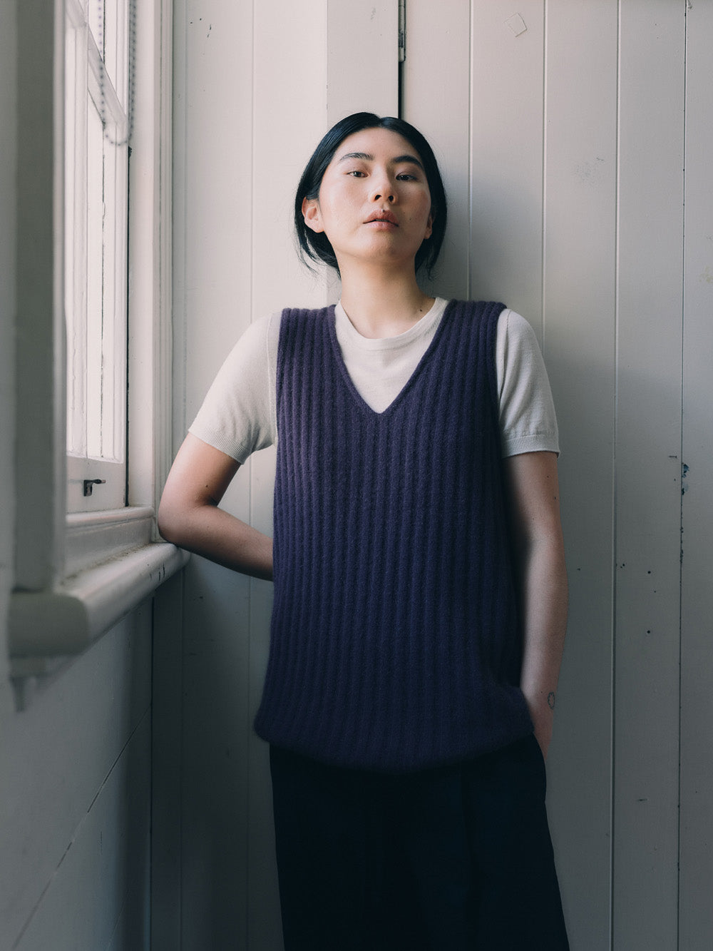 Woman wearing a dark purple knitted vest over a white shirt against a wall.
