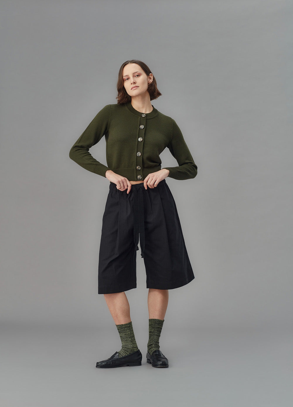Person wearing a green cardigan, black shorts, and green socks on a gray background
