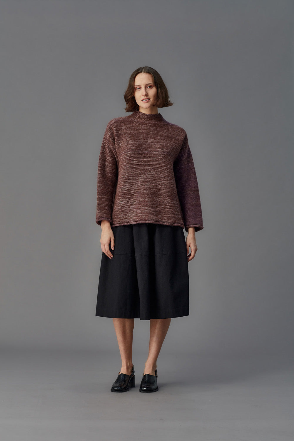 Woman wearing a brown sweater and black skirt against a gray background