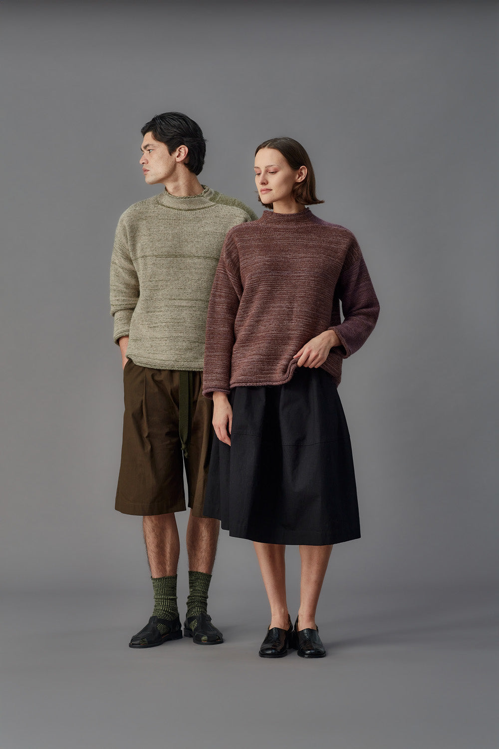 Two people wearing knit sweaters and skirts standing against a gray background