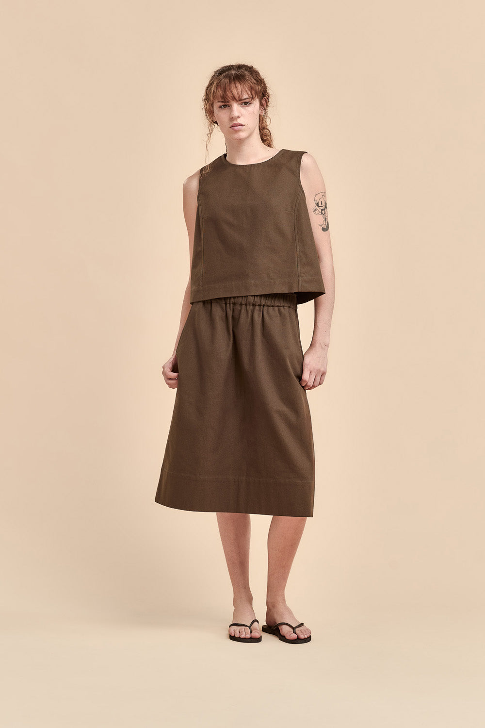 A model stands against a solid beige studio background wearing a sleeveless, cropped brown top and a matching brown midi skirt, paired with black flip-flop sandals.