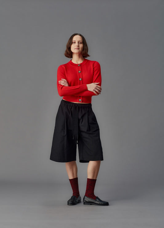 Person wearing a red cardigan, black shorts, and maroon socks on a gray background