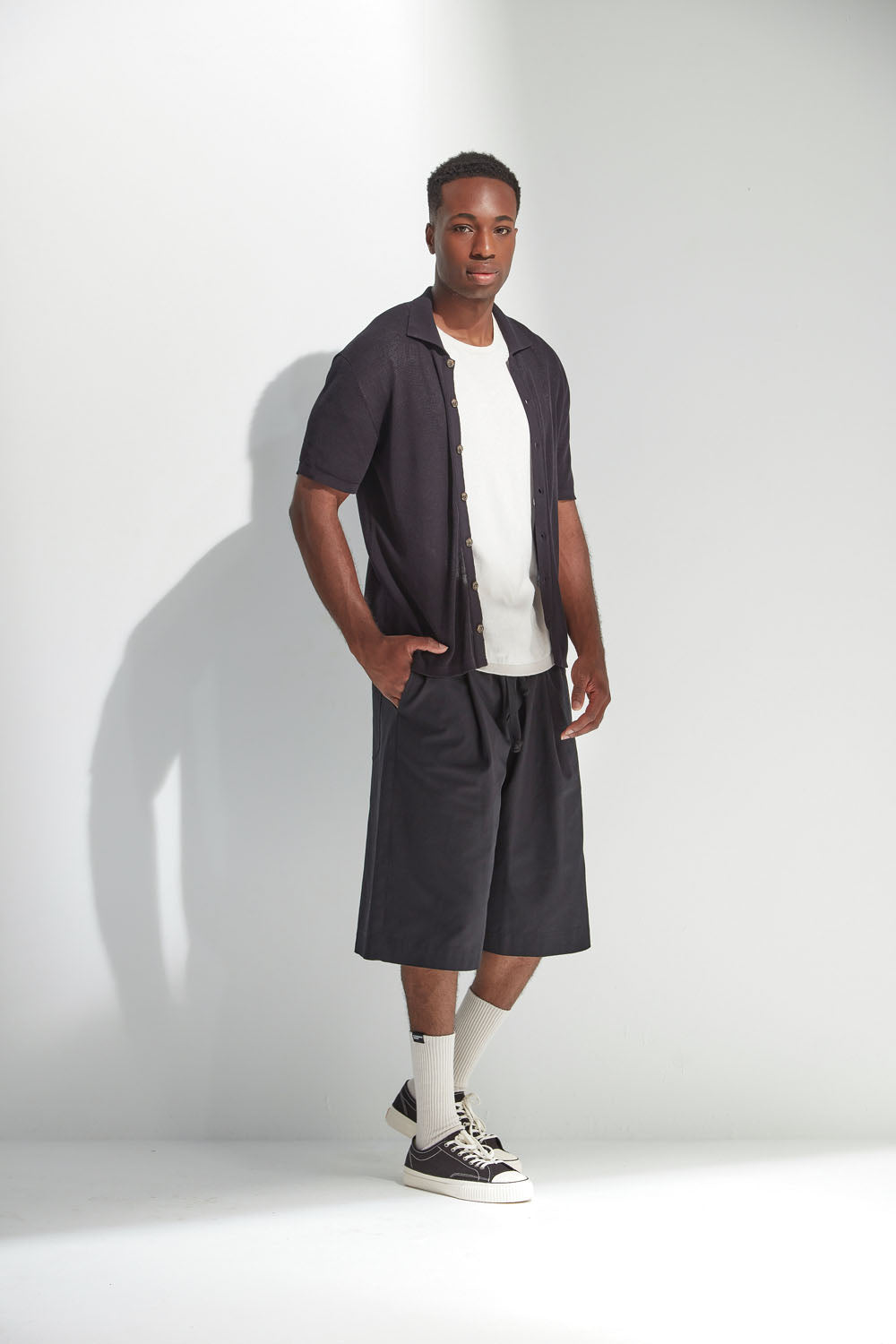 Man wearing a black buttoned shirt with white t-shirt and black shorts against a white background.