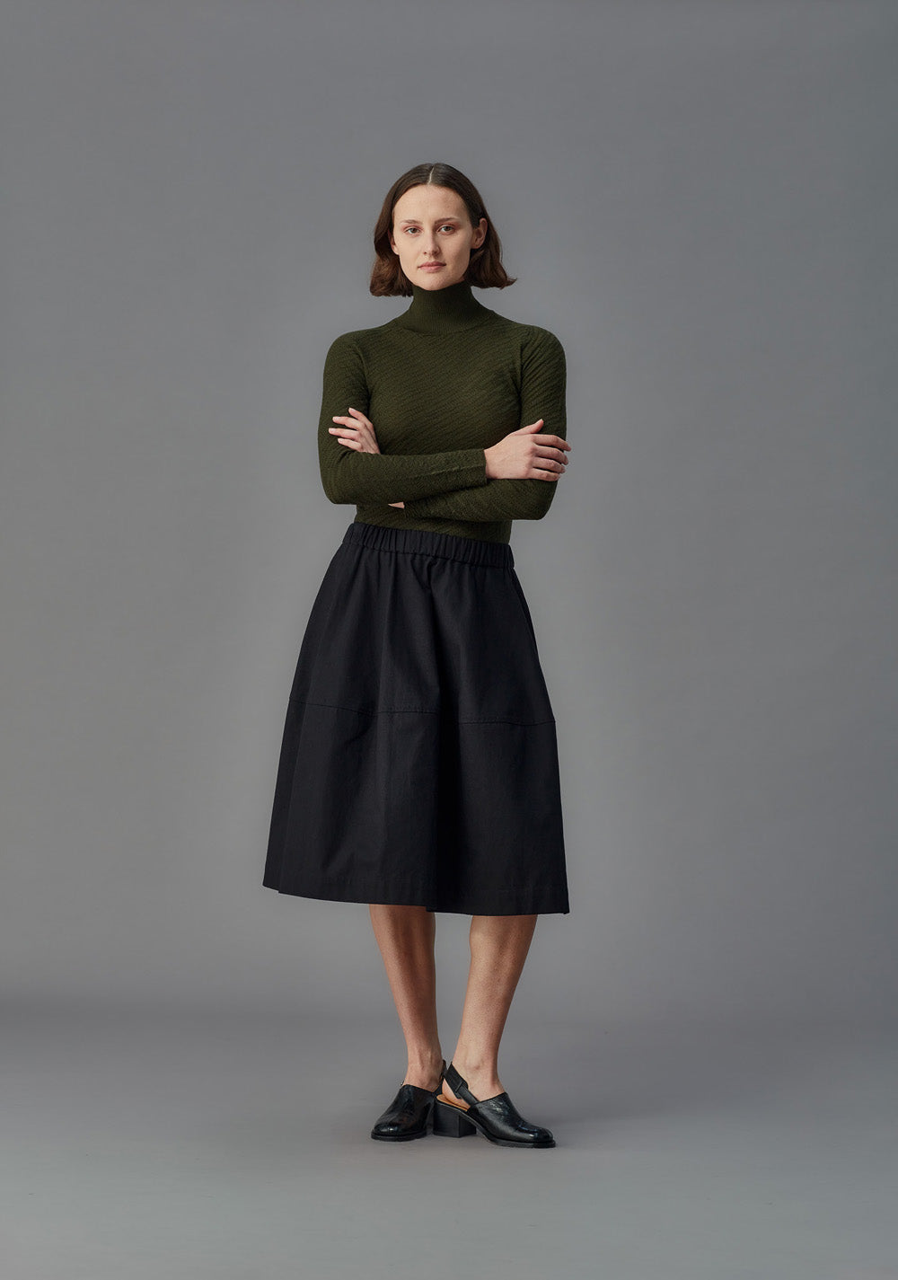 Woman wearing a green turtleneck and black skirt against a gray background