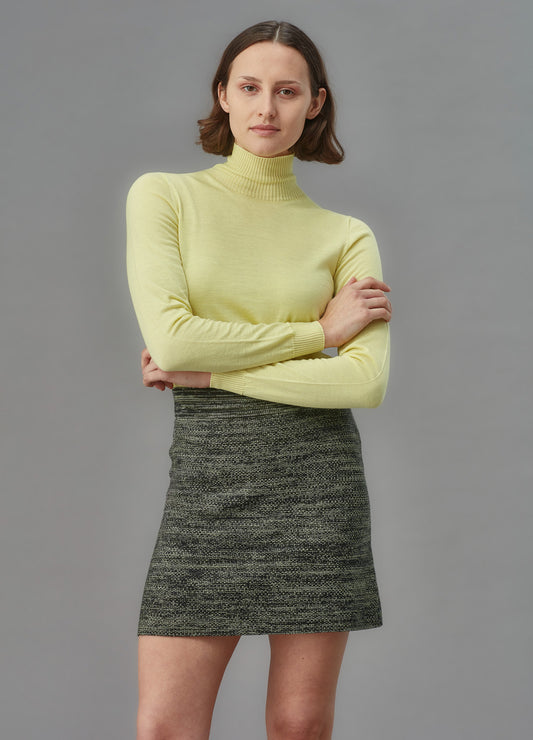 Woman wearing a green knit skirt and light yellow turtleneck sweater against a gray background