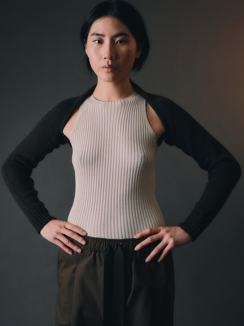 Person wearing black bolero sleeves over a beige ribbed top with dark pants on a dark background