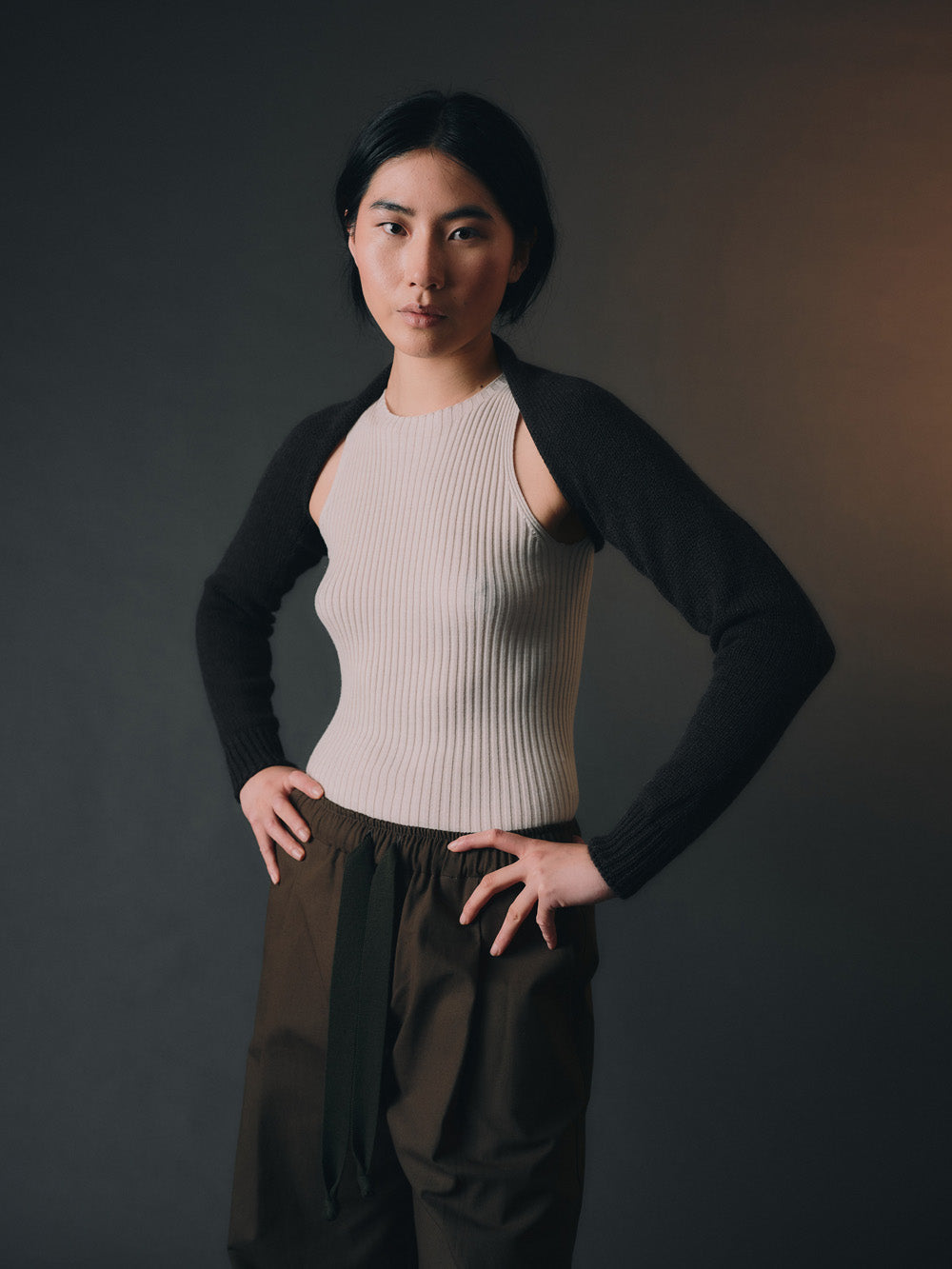 Person wearing black bolero sleeves over a beige ribbed top with dark pants on a dark background