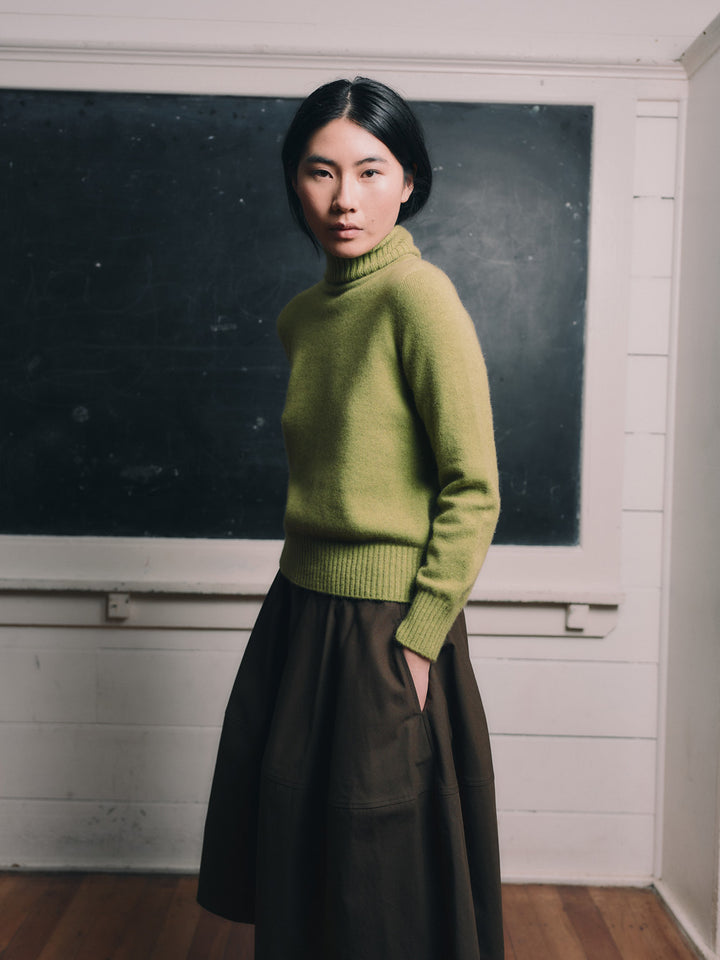 Woman wearing an herb green sweater and brown skirt standing in front of a blackboard.