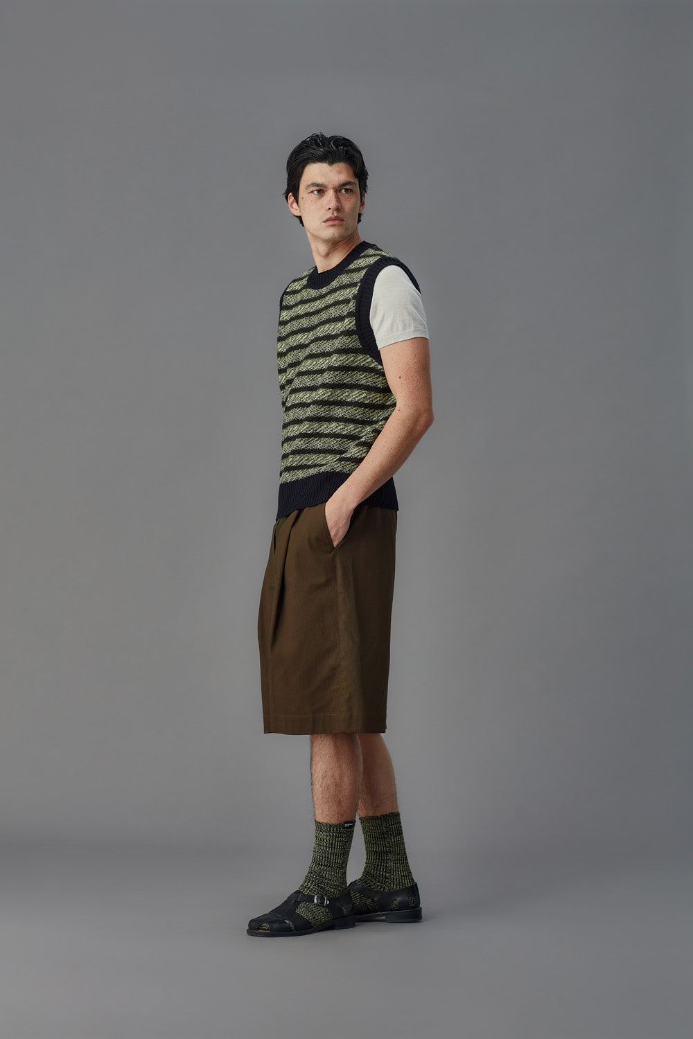 Man wearing a green striped vest, white shirt, brown shorts, and black shoes on a gray background