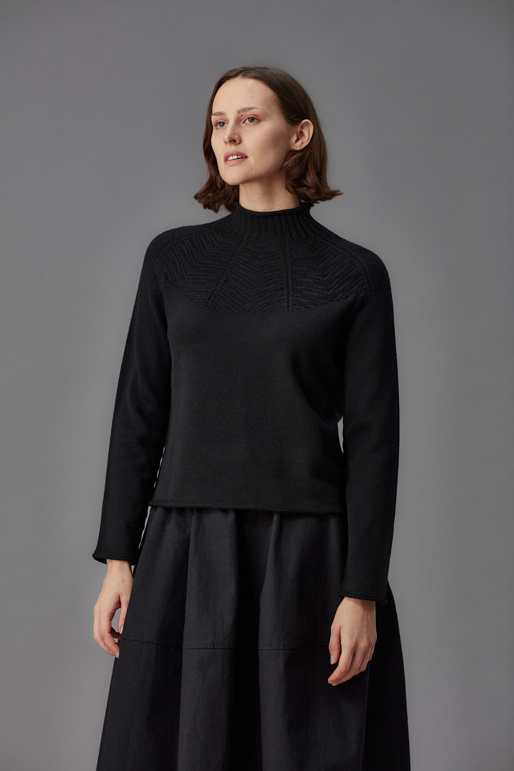 Woman wearing a black mock neck sweater and black skirt against a gray background