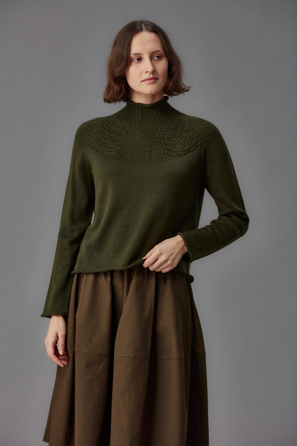 Woman wearing a green mock neck sweater and brown skirt against a gray background