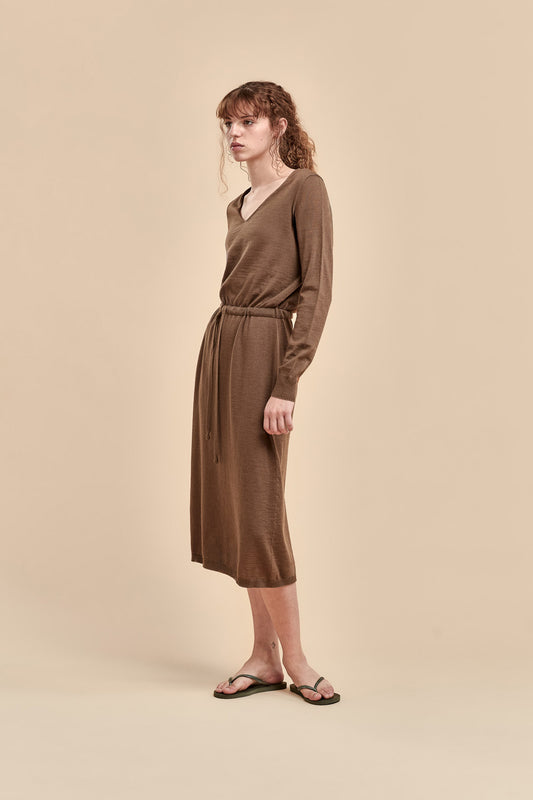 A woman modeling a brown long-sleeve merino dress with a V-neck and a drawstring tie, standing against a solid beige studio background.